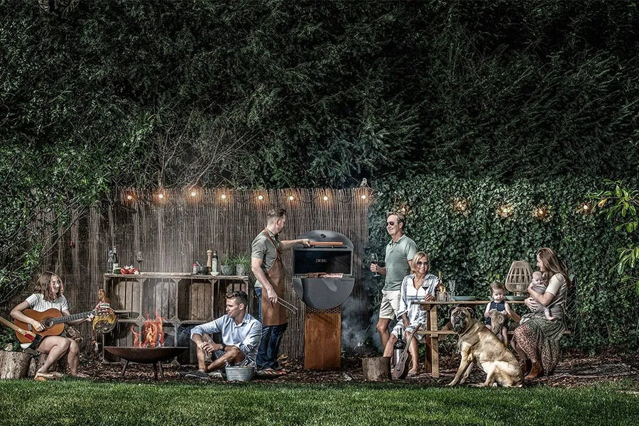 Groep mensen rond een barbecue en tuintafel onder een lichtslinger in de tuin.