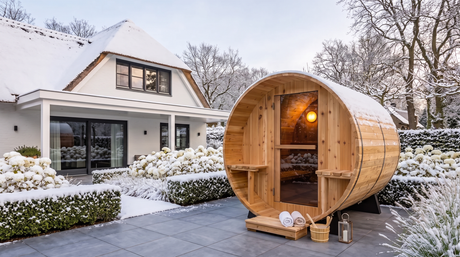 Verlichte houten barrelsauna met trapje in een besneeuwde tuin naast een woning.