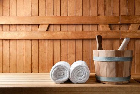 Twee opgerolde handdoeken en een houten emmer op een saunabank.